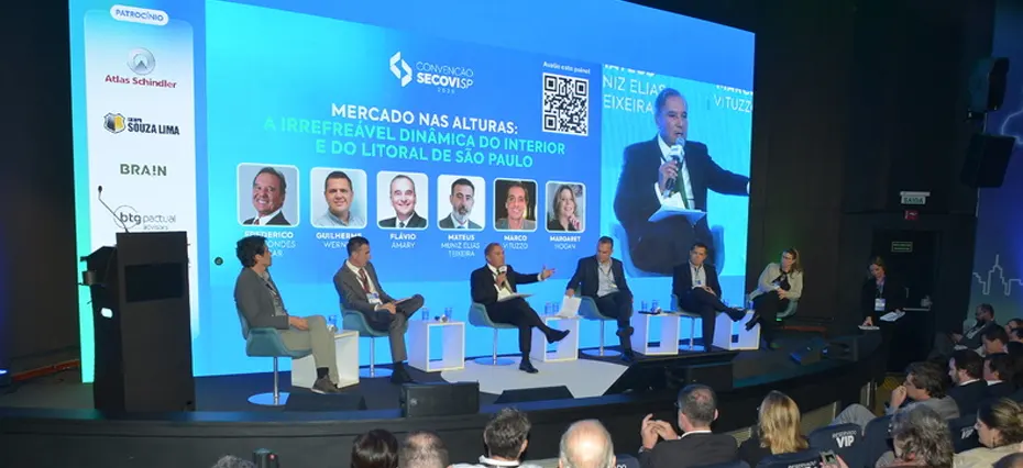 Painel do Secovi que debateu os lançamentos imobiliários em Campinas. Na imagem, sete pessoas estão sentada em cadeiras conversando. No fundo, há uma tela azul com a apresentação do que está sendo abordado.
