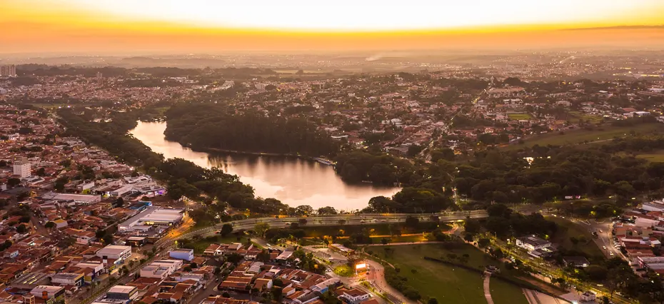Imagem aérea de Campinas, cidade no interior de São Paulo. O céu alaranjado mostra que está no fim da tarde. Há um corpo d'água no canto inferior esquerdo da imagem e muitos prédios no restante do espaço. A foto representa o crescimento dos lançamentos imobiliários em Campinas.